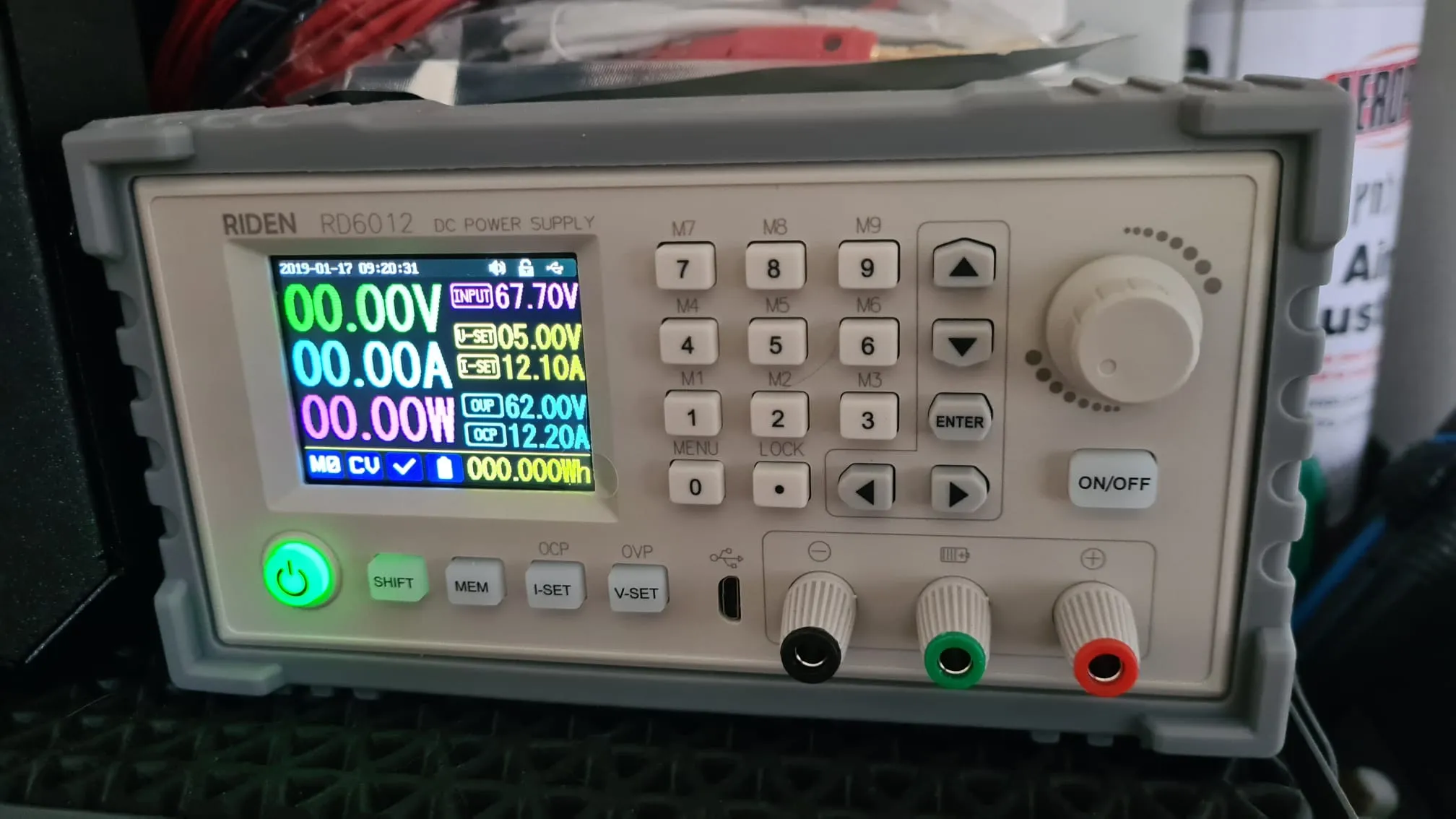 Close-up of a Riden RD6012 bench power supply's front panel, powered on and displaying live voltage, current, and wattage readouts, sitting on a workbench surrounded by wire spools and tools