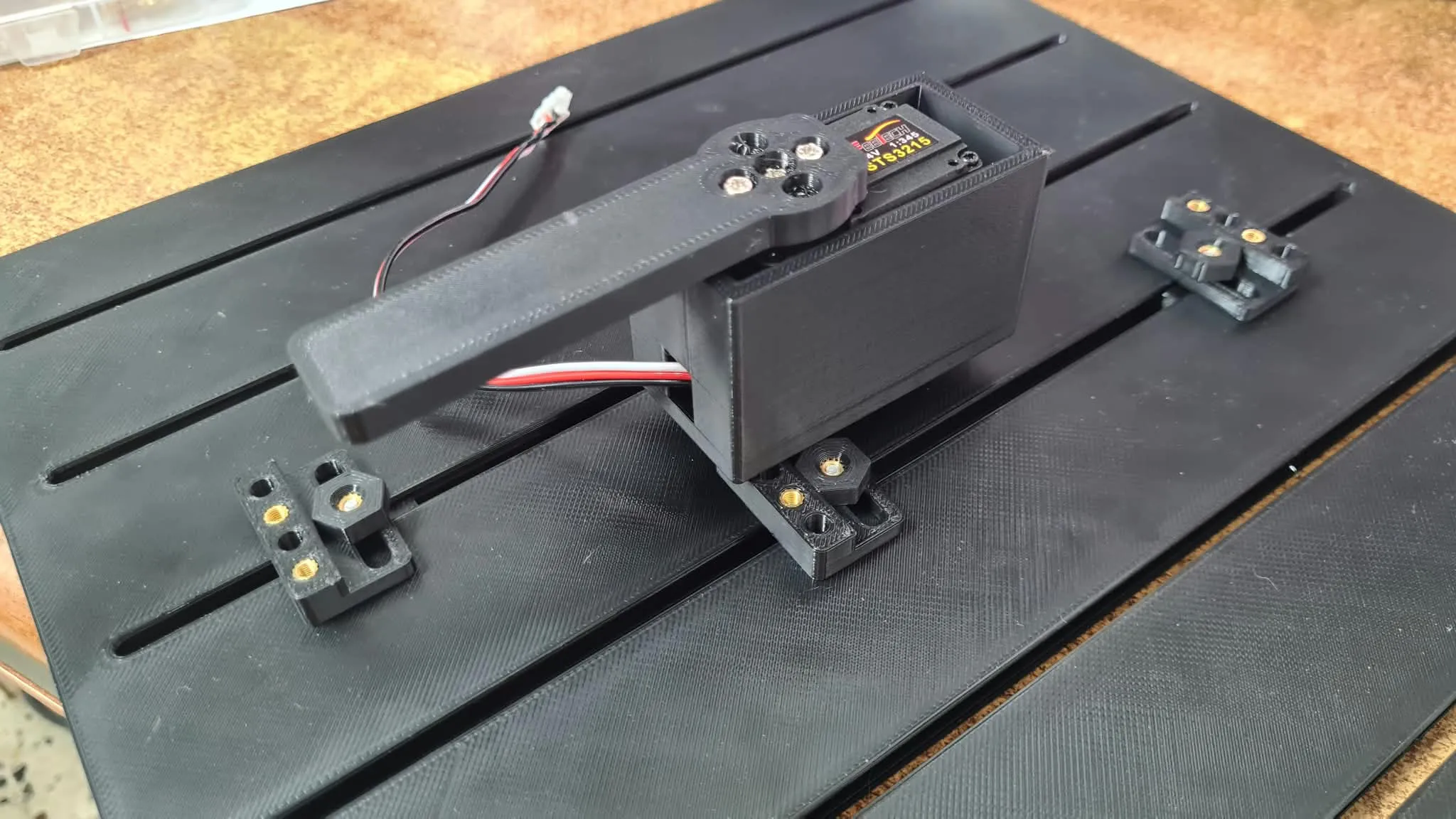 A Feetech STS3215 servo mounted in a black 3D-printed bracket with an output arm attached, wired with a connector cable, sitting on a slotted mounting platform next to a small interposer block