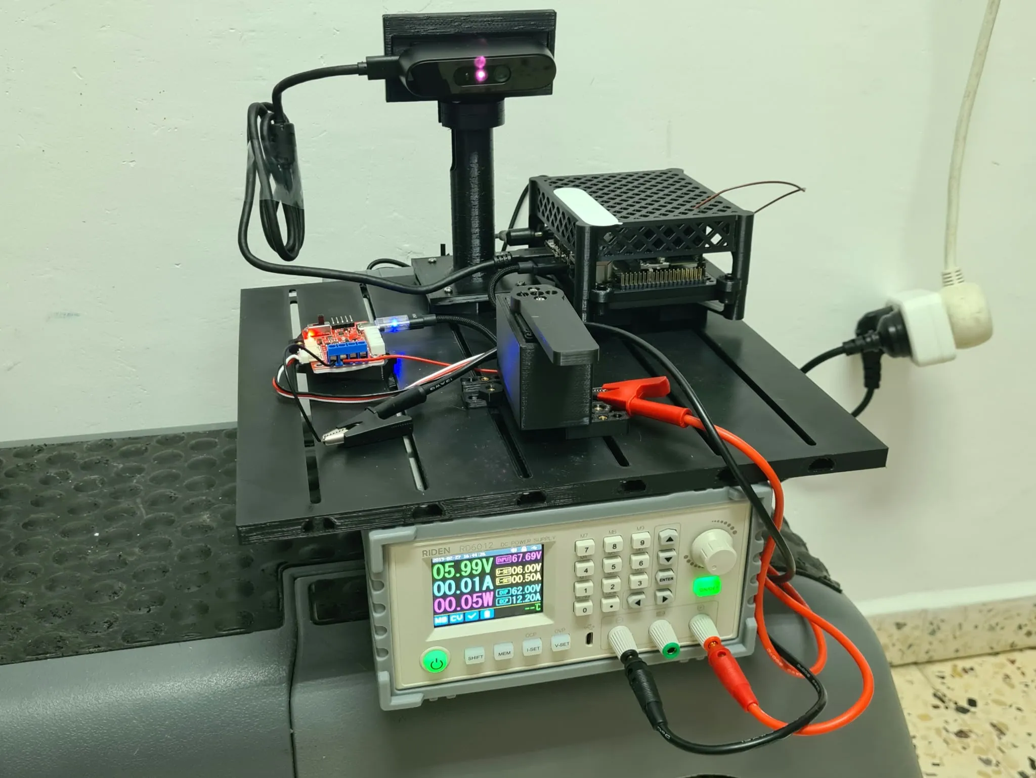 The assembled mounting plate on a bench: a depth camera on a printed tower with its infrared projector lit, a Jetson Orin Nano in its printed lattice enclosure, a small red servo bus board, and a servo in a printed bracket — all sitting above a bench power supply reading 5.99 volts and 0.01 amps