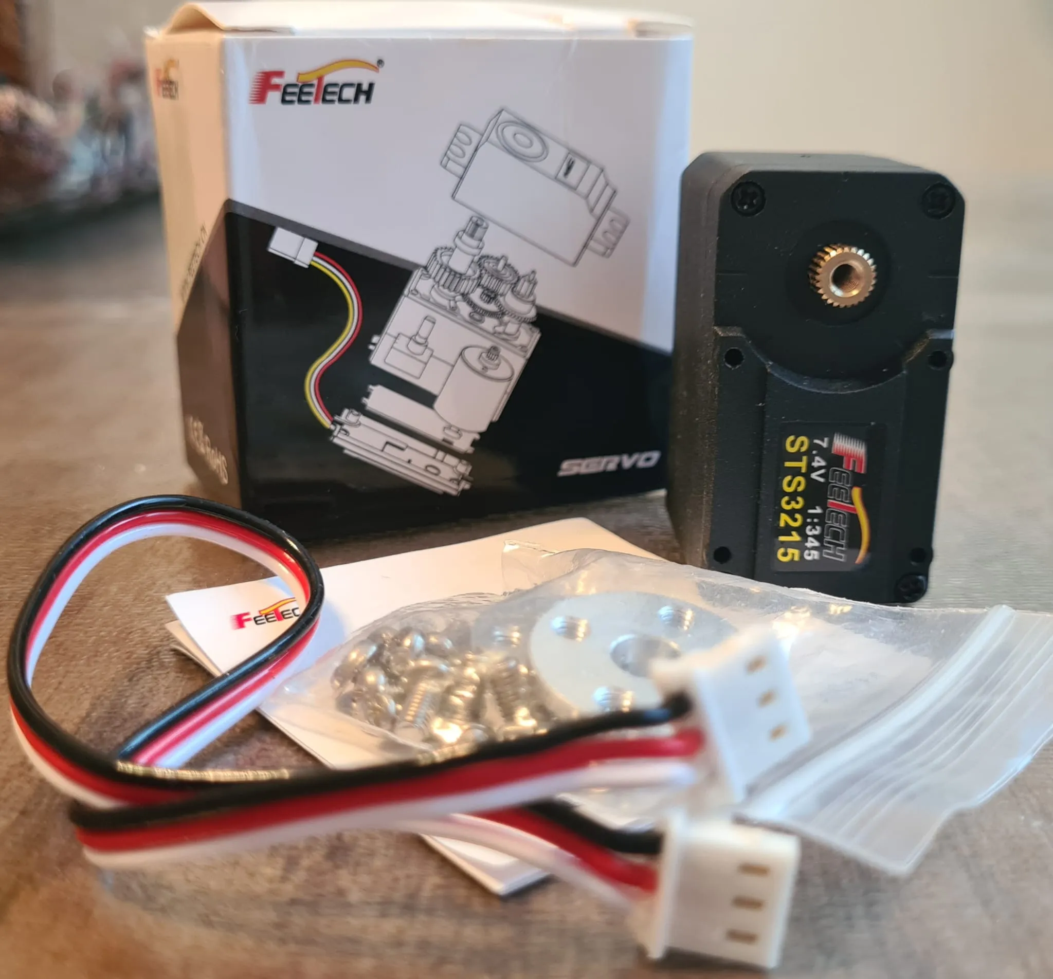 A Feetech STS3215 servo standing next to its own packaging, which shows an exploded-view diagram, with its connector cable, mounting screws, and a small mounting flange laid out on the table in front of it