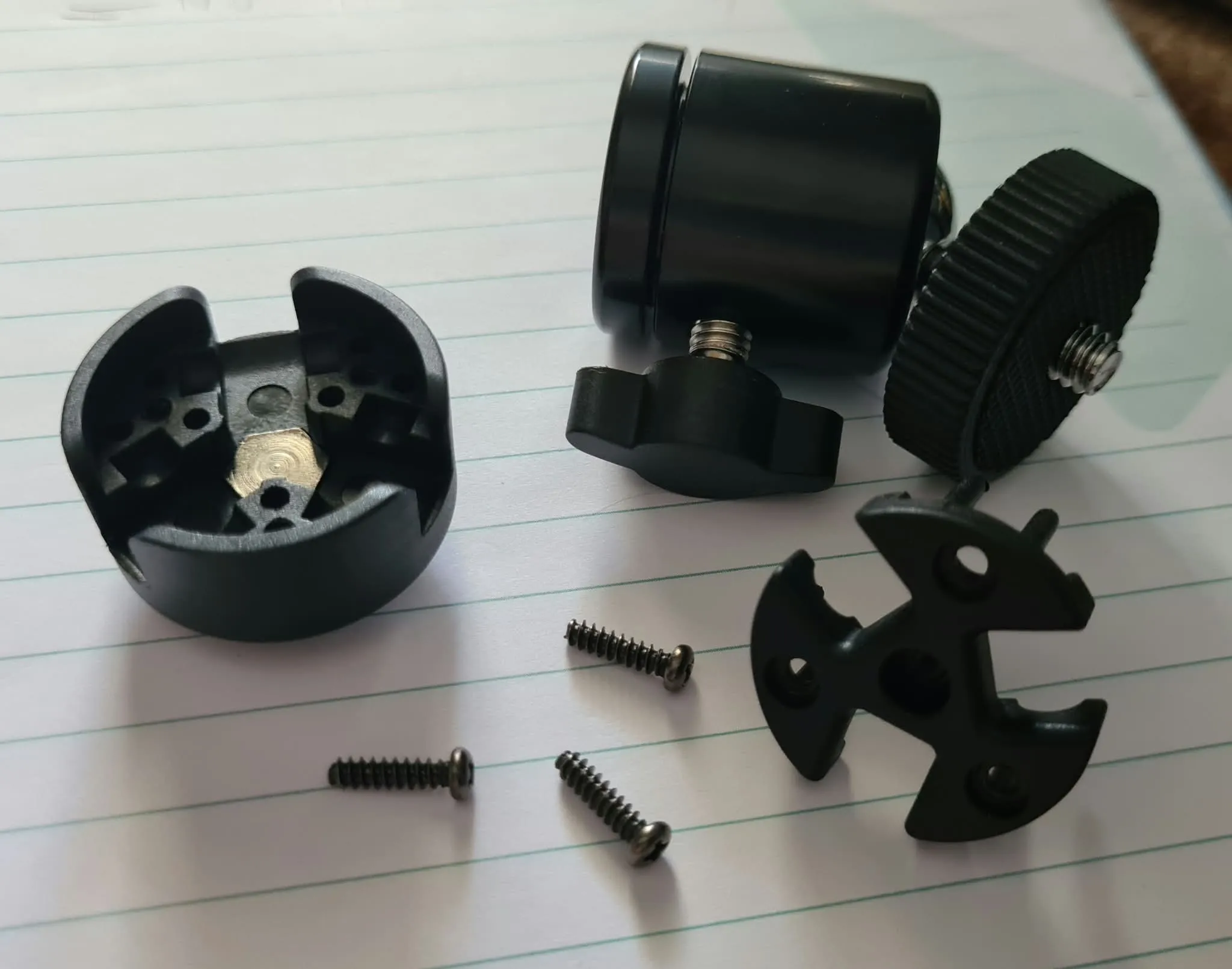A camera tripod's ball head disassembled — ball joint, thumbscrew, collar, clamp plate and three small screws laid out on lined paper